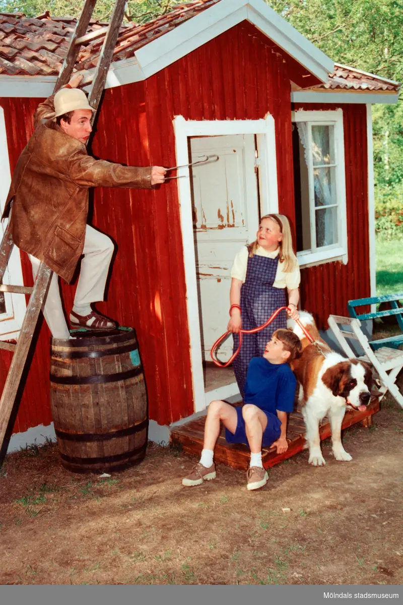 Sommarspelen/barnteaterföreställningen "Tjorven på Saltkråkan" 1996 vid Byxorna i Gunneboskogen. Pappan i familjen (Per Andersson) står på en stege och pekar med en grilltång. Två barn befinner sig vid entrén till ett rött litet hus och flickan håller hunden "Båtsman" i koppel. Relaterade motiv: 2024_0525 - 0533.