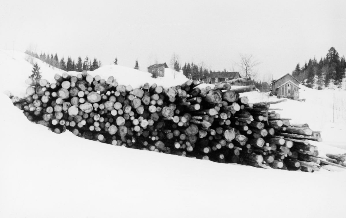 Tømmervelte på snødekt mark i nærheten av Preståa i Enebakk, som har sitt utløp på den sørvestre sida av innsjøen Øyeren.  Fotografiet er tatt mot endeflatene på tømmerstokkene i velta.  Det er snakk om granvirke av varierende dimensjoner og varierende kvalitet, typisk cellulosetømmer.  I dette tilfellet var det råstoff som var innkjøpt til papirproduksjonen ved Greaker Cellulosefabrim AS ved Sarpsborg.  Tømmervelta ligger i et åpent lende.  På en bakkekam i bakgrunnen ser vi husene på et lite gardsbruk.  Bakenfor dette skimter vi litt barskog. 