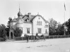 Ett tvåspann har gjort halt framför Asklundska villan i Linköping. Platsen gör det troligt att vi i vagnen ser tobaksfabrikören Ernst Asklund vid hustrun och parets yngsta barn. Villan hade makarna låtit uppföra 1894, nu skrev man 1902.