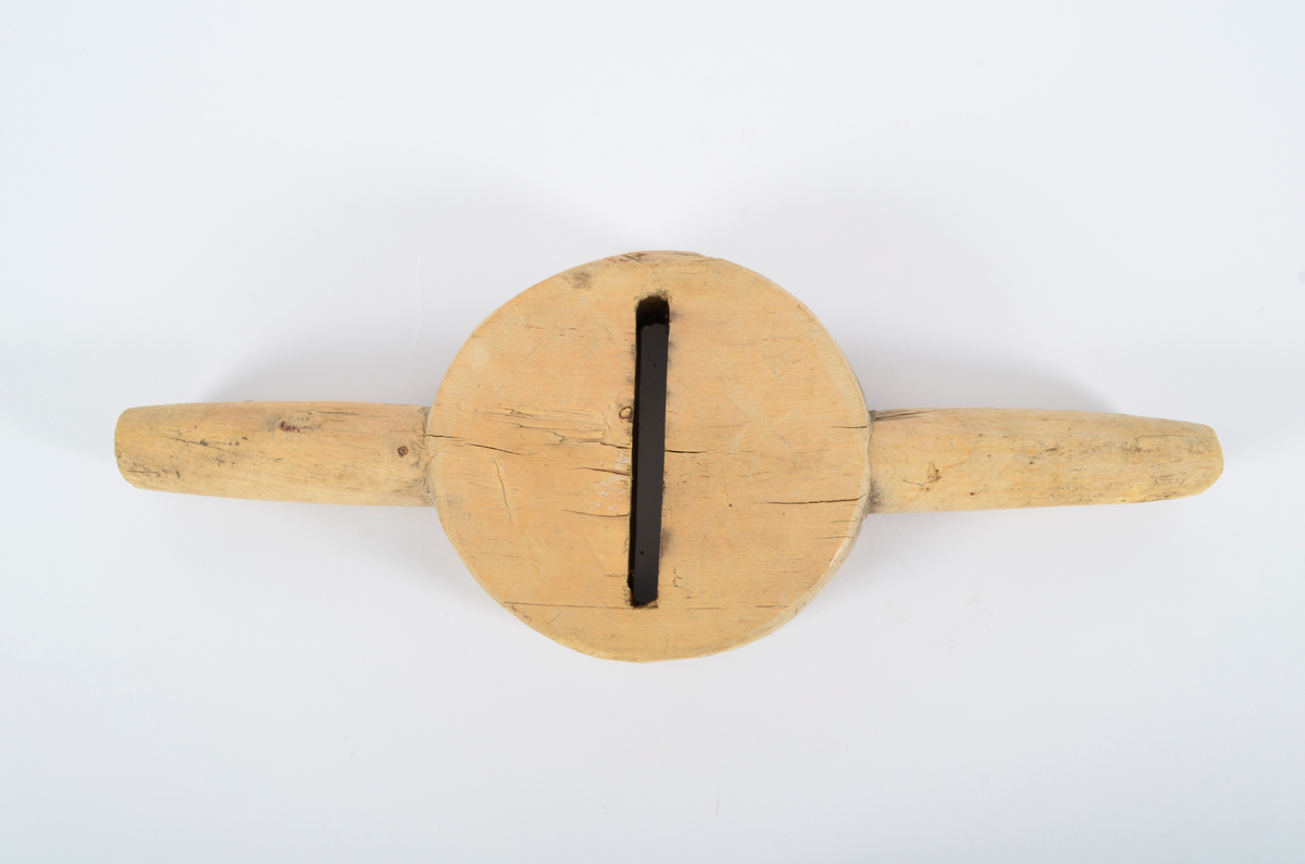 Består av sirkulær kloss med gjennombrutt smal fure i sentrum, ca. 12 cm lang. Horisontalt på furen, to runde håndtak. Smalner utover. Umalt.