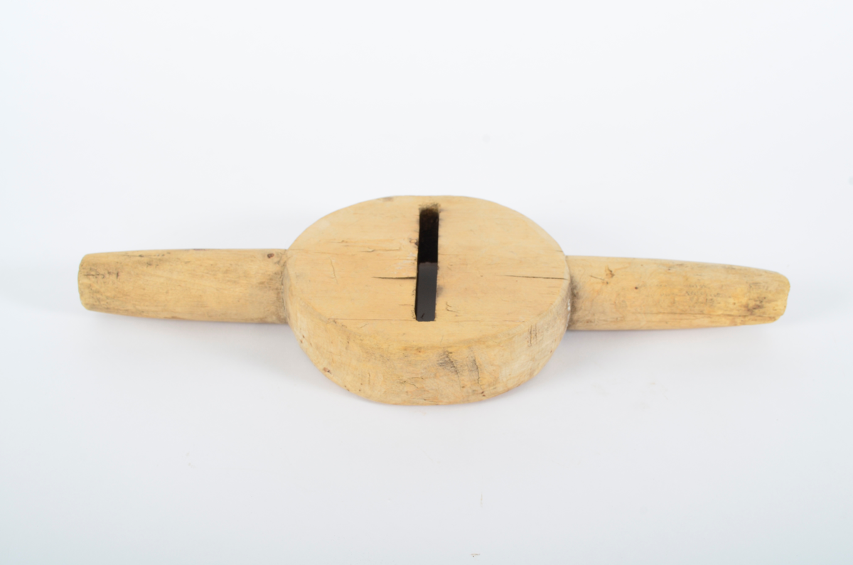 Består av sirkulær kloss med gjennombrutt smal fure i sentrum, ca. 12 cm lang. Horisontalt på furen, to runde håndtak. Smalner utover. Umalt.