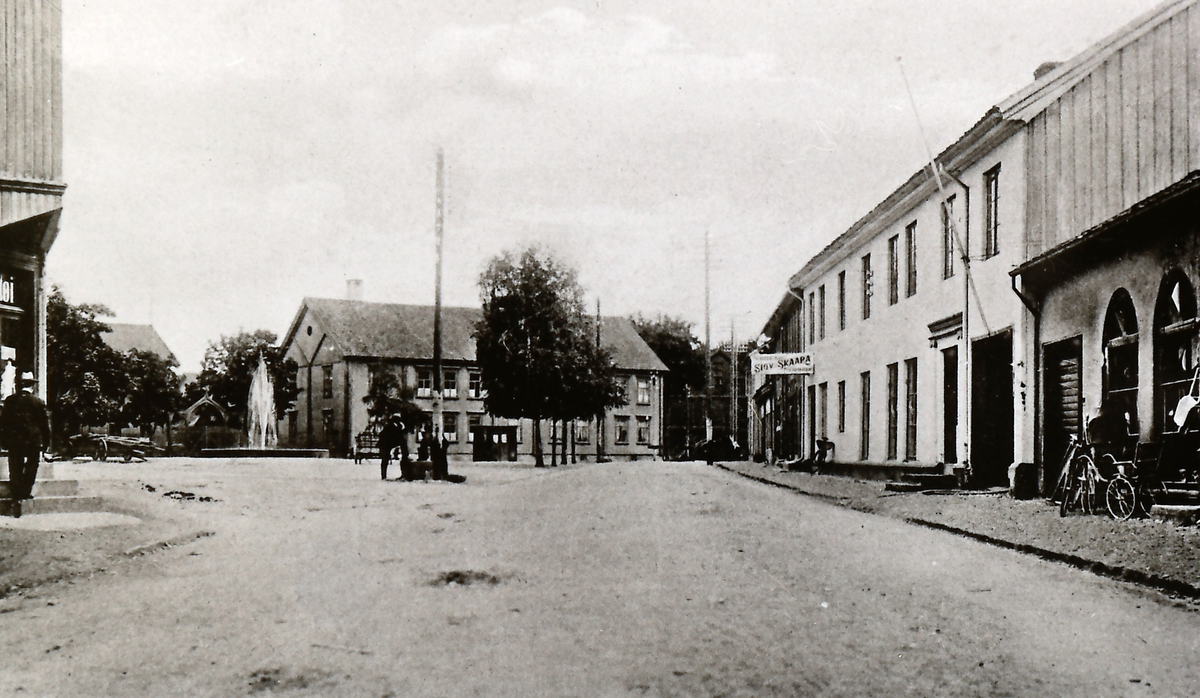 Tråkka (Nymoens torg) med fontenen.
Elektrisitetsverket ( startet 1911) har begynt å reise stolper. Bakerst Kongsberg Middelskole, der den fine rokokkodøra er flyttet inn mot gården.