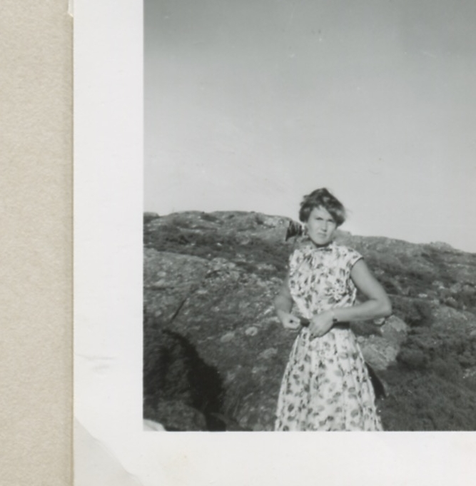 En ung okänd kvinna, klädd i en blommig klänning, står framför några bergsknallar, Backen eller Högen 1960-tal.