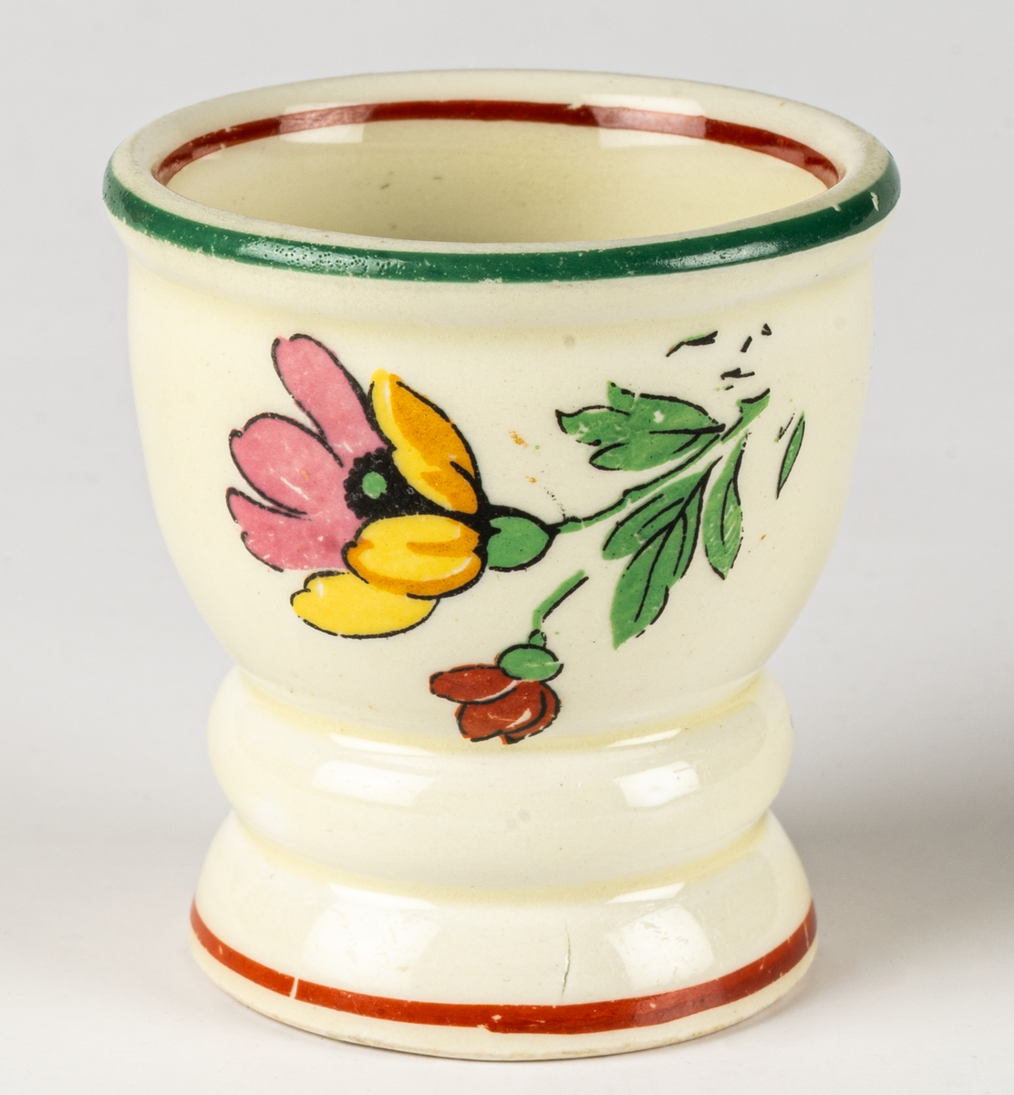 Äggkopp i flintgods, modell B, dekor Carmen. Dekoren ritad av Eugen Trost, tillverkad 1933–45. Carmen är en kromotryckt dekor med blommor på ljust gul glasyr med kantränder i rött och grönt.