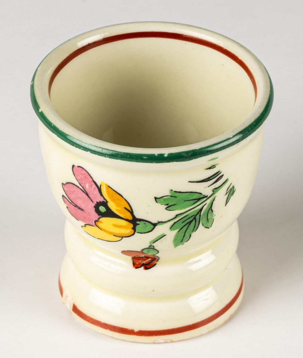 Äggkopp i flintgods, modell B, dekor Carmen. Dekoren ritad av Eugen Trost, tillverkad 1933–45. Carmen är en kromotryckt dekor med blommor på ljust gul glasyr med kantränder i rött och grönt.