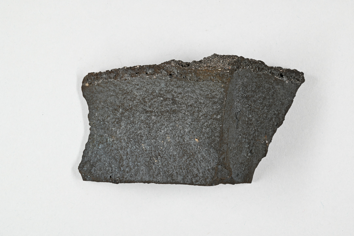 Materialprov, grå-svart. Tillhörande lapp med notering: "1:sta skärsten från Pb-smältning i Kafveltorp, Juli 1902, J. Petrén".