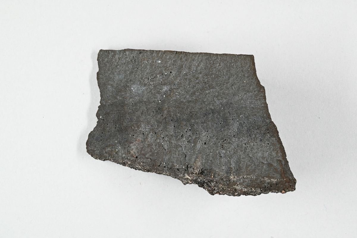 Materialprov, grå-svart. Tillhörande lapp med notering: "1:sta skärsten från Pb-smältning i Kafveltorp, Juli 1902, J. Petrén".