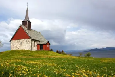 Edøy kirke på Smøla, steinbygning med hvitkalkede murer, rødmalt treverk. Blomstereng og utsikt utover leia.