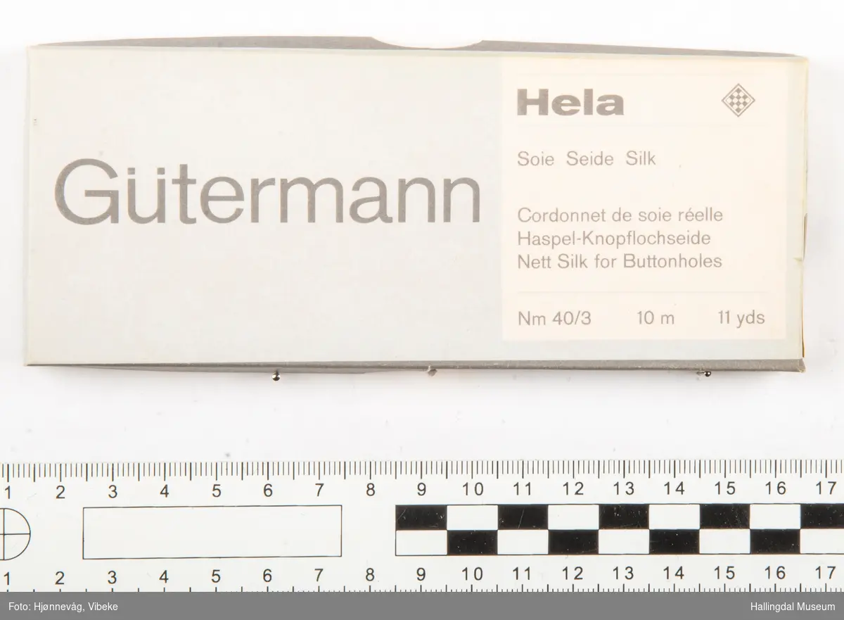 Eske som inneholder 7 silkespoler. Esken er fra "Gutermann". På den ene kortsiden står nummeret "752". Det er en firkantet tegning på samme kortside og det står "Hela 10 m". På toppen av esken står følgende: "Sore Seide Silk Cordonnet de sore rèelle Haspel-Knopflochseide Nett Silk for Buttomholles. NM 40/3 10 m 11 yds.