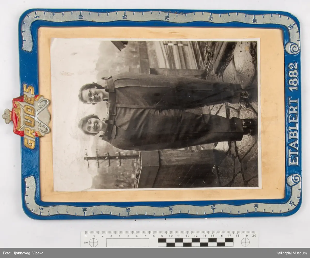 Blå bilderamme i metall "Grudes etablert 1882". Bilde av 2 damer i hvert sitt ben på en stor skreddersydd bukse.