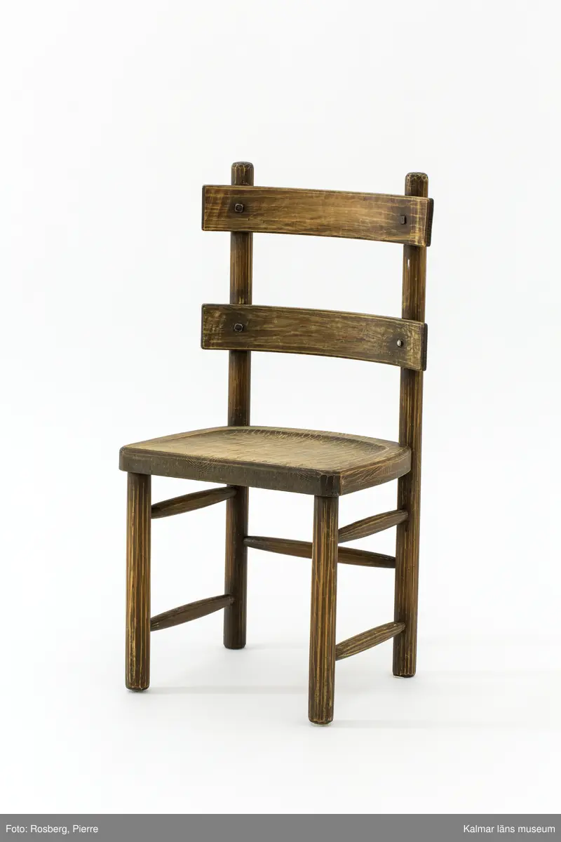 KLM 46378:3. Stol. Ingår i möbelserien "Sandhamn". Av furu med brunbetsad yta som senare även slipats. Skålad sits. Ryggen består av två ribbor fastsatta med smidda järnspikar, ena spikhuvudet saknas. Under stolssitsen en rektangulär metallbricka med text: AB NORDISKA KOMPANIET R 33845- MÖB.