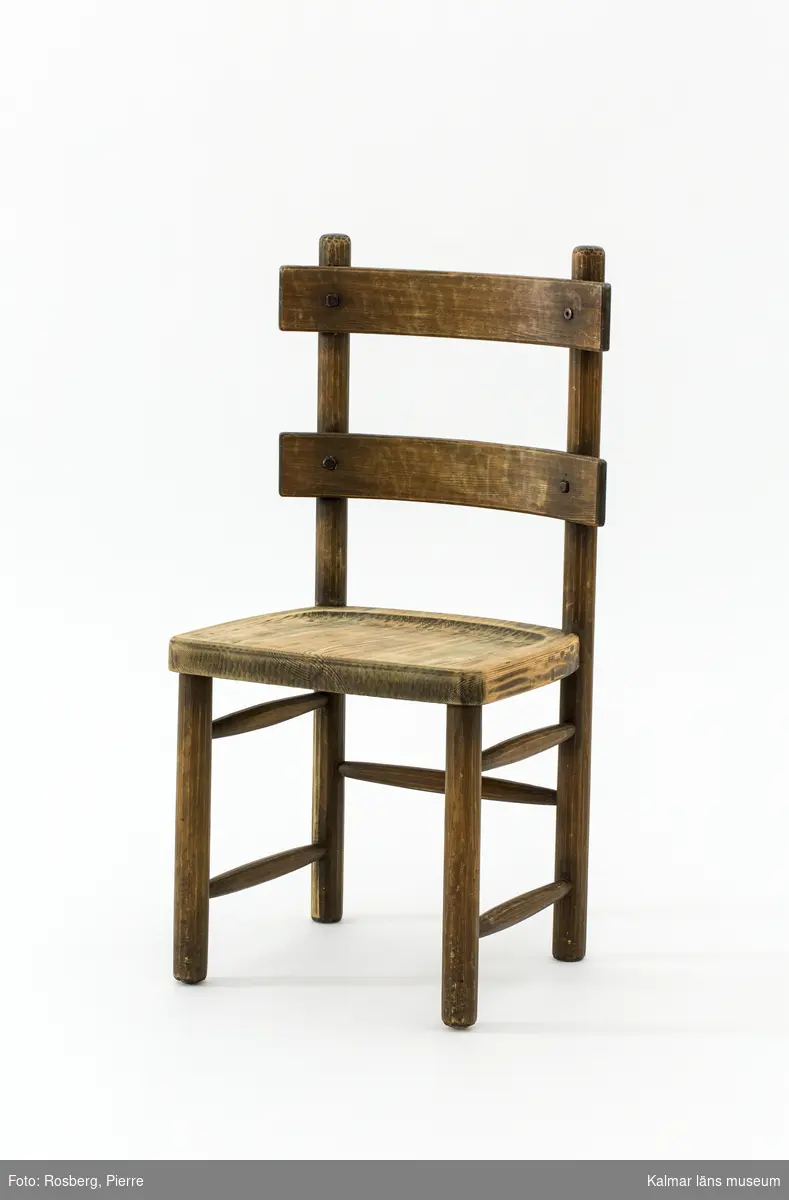 KLM 46378:5. Stol. Ingår i möbelserien "Sandhamn". Av furu med brunbetsad yta som senare även slipats. Skålad sits. Ryggen består av två ribbor fastsatta med smidda järnspikar, ena spikhuvudet saknas. Under stolssitsen märken efter en rektangulär metallbricka.