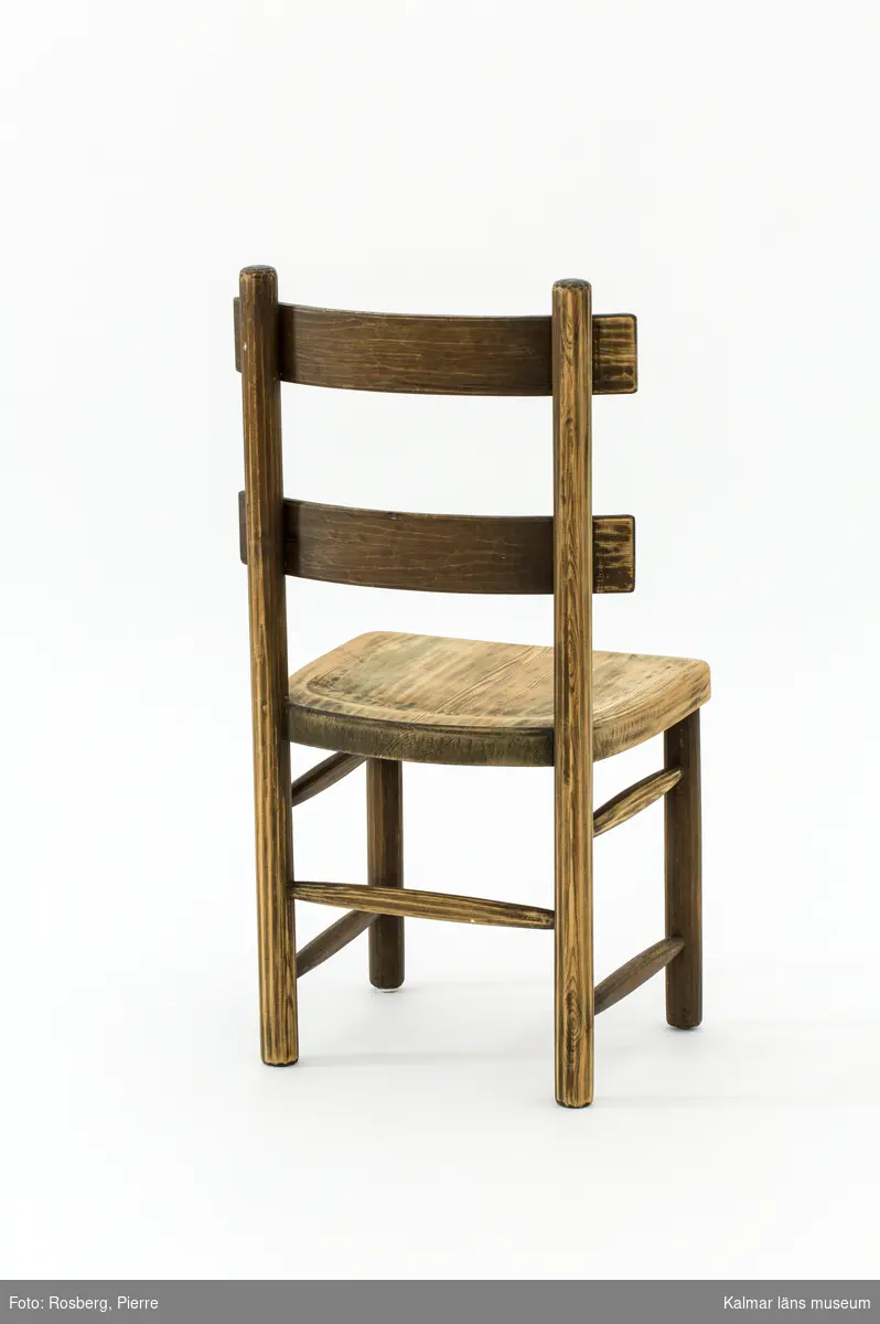 KLM 46378:5. Stol. Ingår i möbelserien "Sandhamn". Av furu med brunbetsad yta som senare även slipats. Skålad sits. Ryggen består av två ribbor fastsatta med smidda järnspikar, ena spikhuvudet saknas. Under stolssitsen märken efter en rektangulär metallbricka.