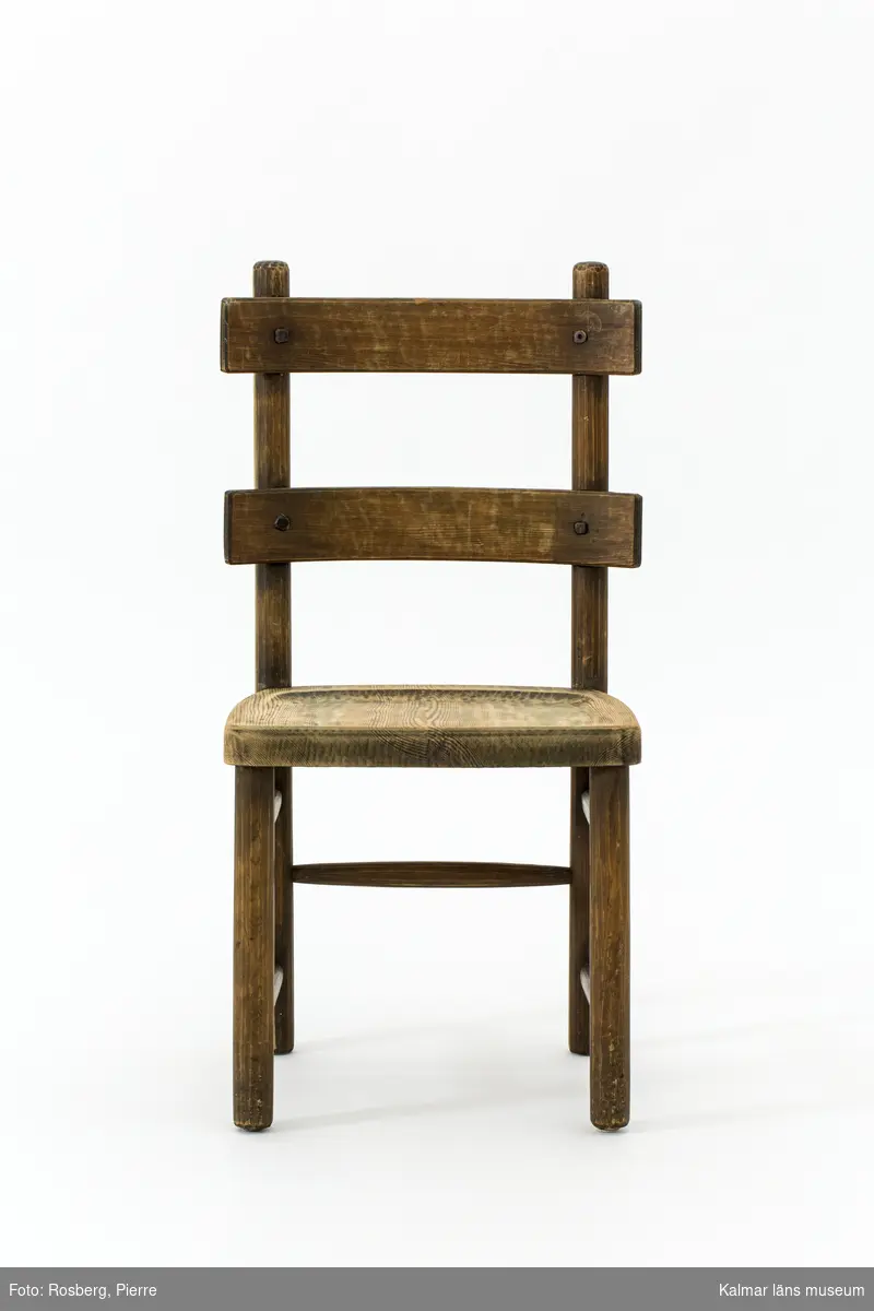 KLM 46378:5. Stol. Ingår i möbelserien "Sandhamn". Av furu med brunbetsad yta som senare även slipats. Skålad sits. Ryggen består av två ribbor fastsatta med smidda järnspikar, ena spikhuvudet saknas. Under stolssitsen märken efter en rektangulär metallbricka.