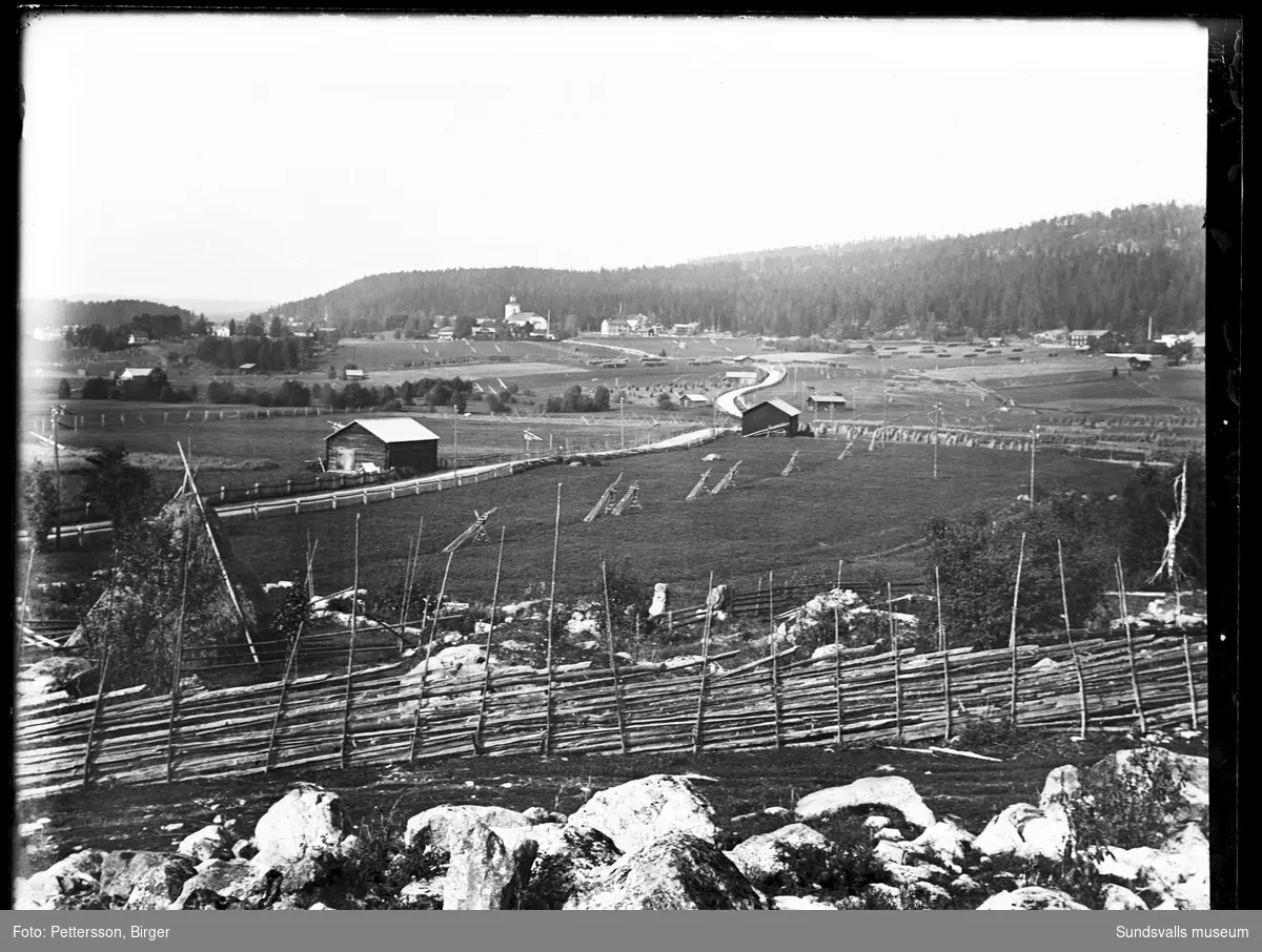 Landsbygdsvy med jordbrukslandskap, hagar, lador och gårdar samt längre bort en kyrka.