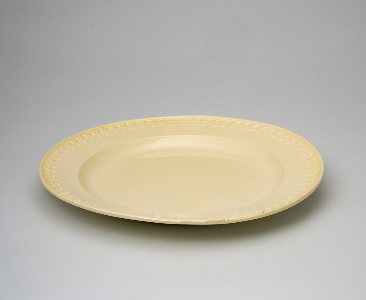 En flat tallerken av glasert steintøy (creamware). Den er hvitgul på farge (ensfarget). Flaten på midten er 16,5cm i diameter. Langs kanten er det tungekant. Innenfor tungekanten er det støpt dekor i form av en hjertebord med stiliserte blomster/bladverk. På undersiden er det dypstempel i form av fire små hjerteformede prikker i klase (utgjør en sirkerl) som er et pottemakermerke.