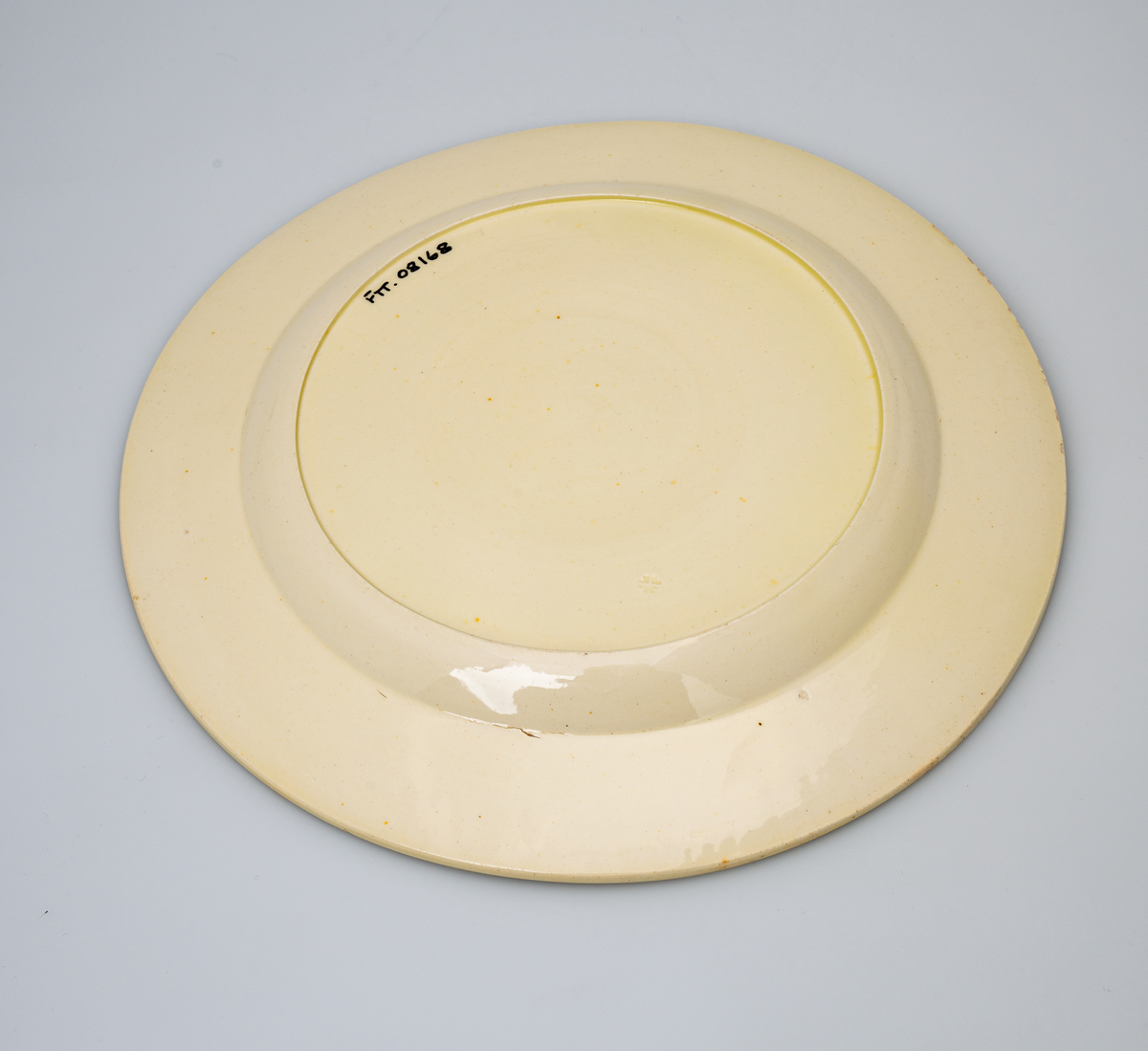 En flat tallerken av glasert steintøy (creamware). Den er hvitgul på farge (ensfarget). Flaten på midten er 16,5cm i diameter. Langs kanten er det tungekant. Innenfor tungekanten er det støpt dekor i form av en hjertebord med stiliserte blomster/bladverk. På undersiden er det dypstempel i form av fire små hjerteformede prikker i klase (utgjør en sirkerl) som er et pottemakermerke.