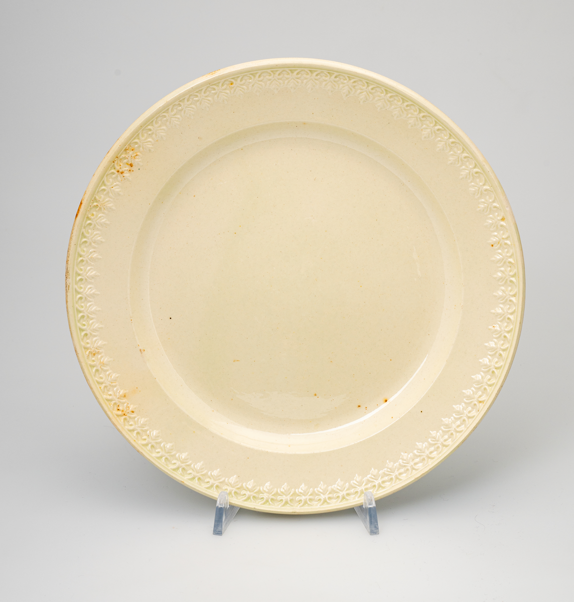 En flat tallerken av glasert steintøy (creamware). Den er hvitgul på farge (ensfarget). Flaten på midten er 16,5cm i diameter. Langs kanten er det tungekant. Innenfor tungekanten er det støpt dekor i form av en hjertebord med stiliserte blomster/bladverk. På undersiden er det dypstempel i form av fire små ruter/prikker i klase som er et pottemakermerke.