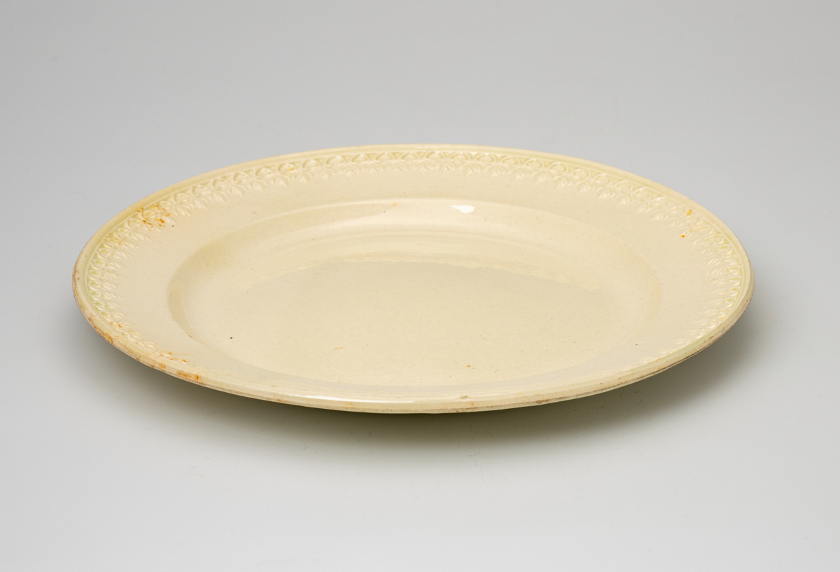 En flat tallerken av glasert steintøy (creamware). Den er hvitgul på farge (ensfarget). Flaten på midten er 16,5cm i diameter. Langs kanten er det tungekant. Innenfor tungekanten er det støpt dekor i form av en hjertebord med stiliserte blomster/bladverk. På undersiden er det dypstempel i form av fire små ruter/prikker i klase som er et pottemakermerke.