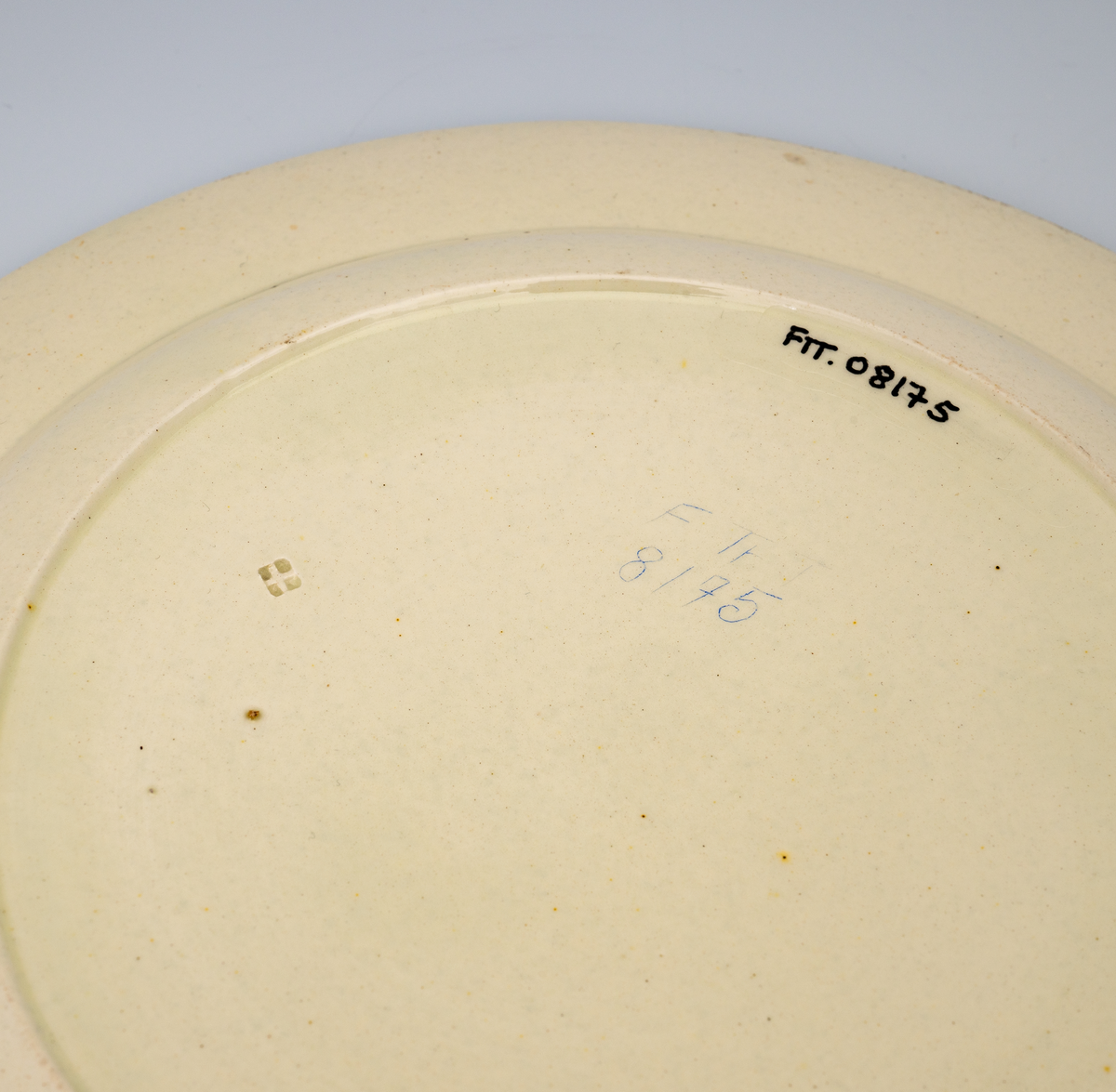 En flat tallerken av glasert steintøy (creamware). Den er hvitgul på farge (ensfarget). Flaten på midten er 16,5cm i diameter. Langs kanten er det tungekant. Innenfor tungekanten er det støpt dekor i form av en hjertebord med stiliserte blomster/bladverk. På undersiden er det dypstempel i form av fire små ruter/prikker i klase som er et pottemakermerke.