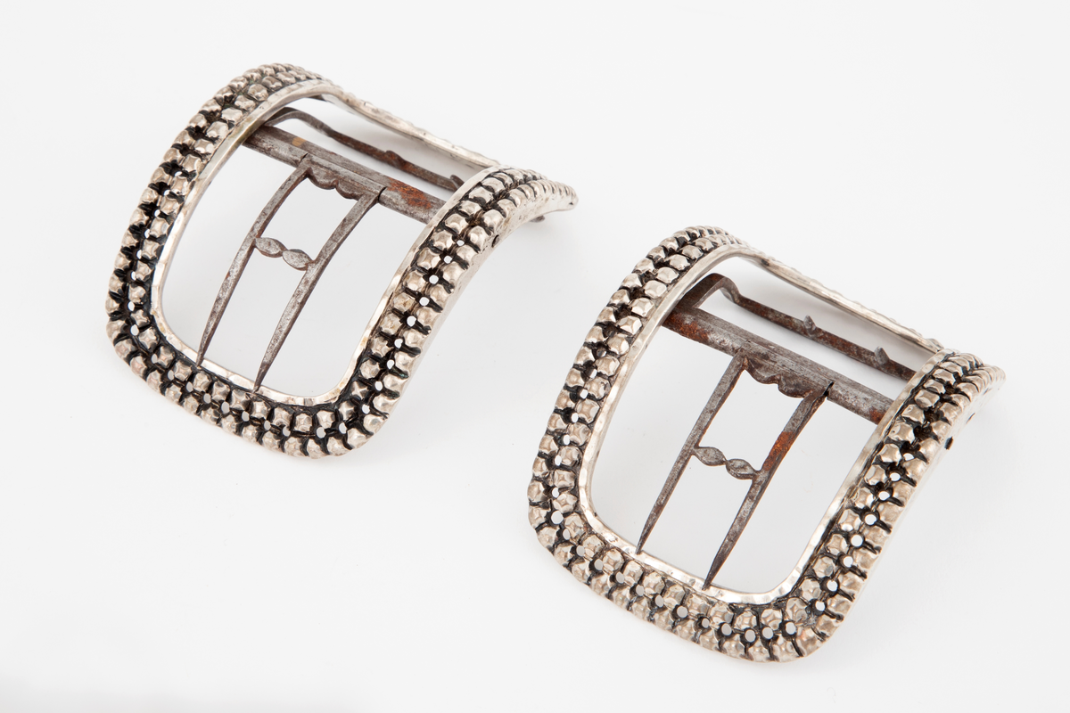 Skospenner støpt i sølv og siselert. Krummet, avlang rammeform med lett buede kortsider. Dekorert med to rader kantede "knopper", skilt med en rad små, gjennombrutte hull. Stolpe med dobbel torn i form av to parallelle tinner og feste med 2 små, faste torner, alt av jern.