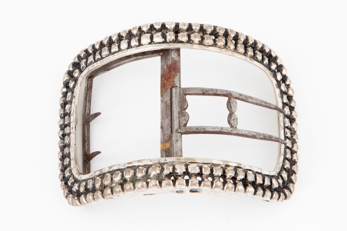 Skospenner støpt i sølv og siselert. Krummet, avlang rammeform med lett buede kortsider. Dekorert med to rader kantede "knopper", skilt med en rad små, gjennombrutte hull. Stolpe med dobbel torn i form av to parallelle tinner og feste med 2 små, faste torner, alt av jern.