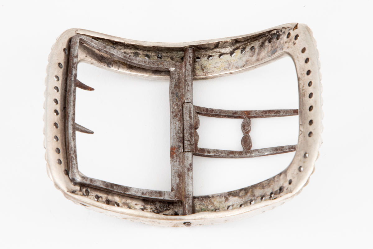 Skospenner støpt i sølv og siselert. Krummet, avlang rammeform med lett buede kortsider. Dekorert med to rader kantede "knopper", skilt med en rad små, gjennombrutte hull. Stolpe med dobbel torn i form av to parallelle tinner og feste med 2 små, faste torner, alt av jern.