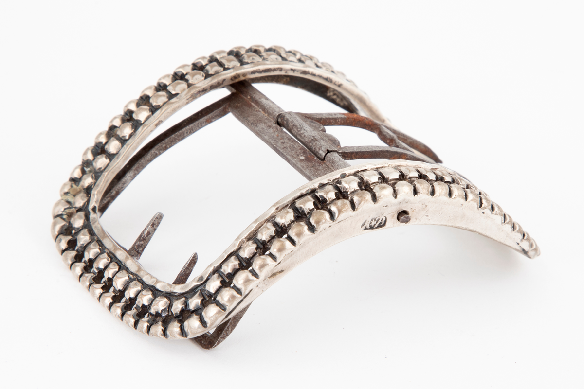 Skospenner støpt i sølv og siselert. Krummet, avlang rammeform med lett buede kortsider. Dekorert med to rader kantede "knopper", skilt med en rad små, gjennombrutte hull. Stolpe med dobbel torn i form av to parallelle tinner og feste med 2 små, faste torner, alt av jern.