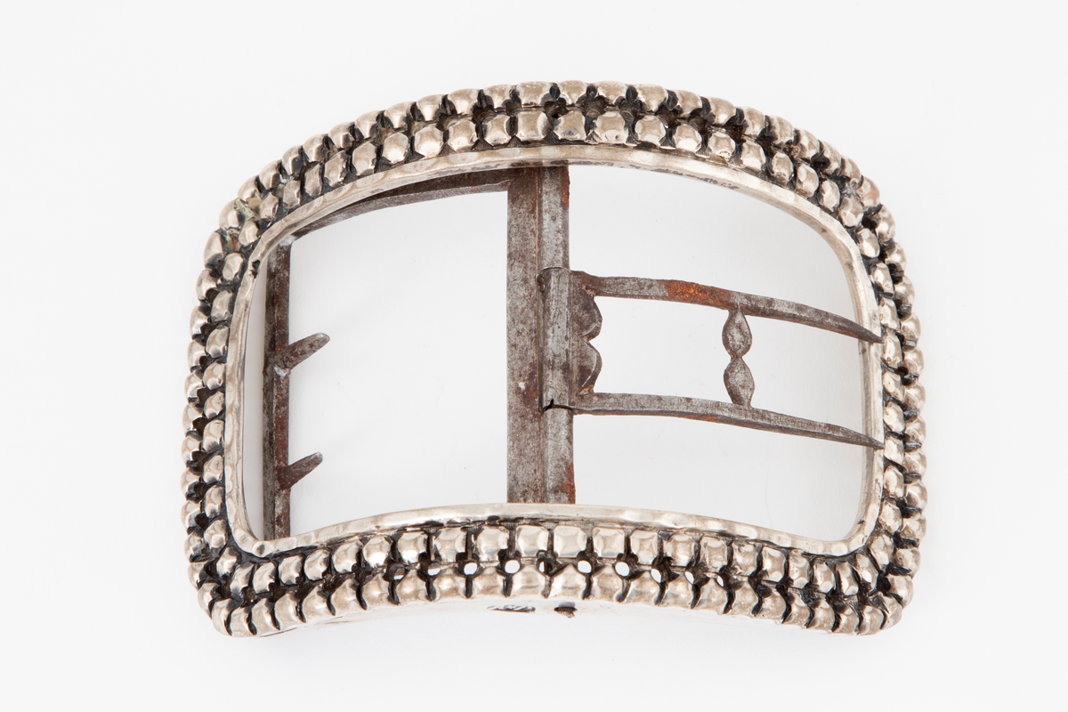 Skospenner støpt i sølv og siselert. Krummet, avlang rammeform med lett buede kortsider. Dekorert med to rader kantede "knopper", skilt med en rad små, gjennombrutte hull. Stolpe med dobbel torn i form av to parallelle tinner og feste med 2 små, faste torner, alt av jern.