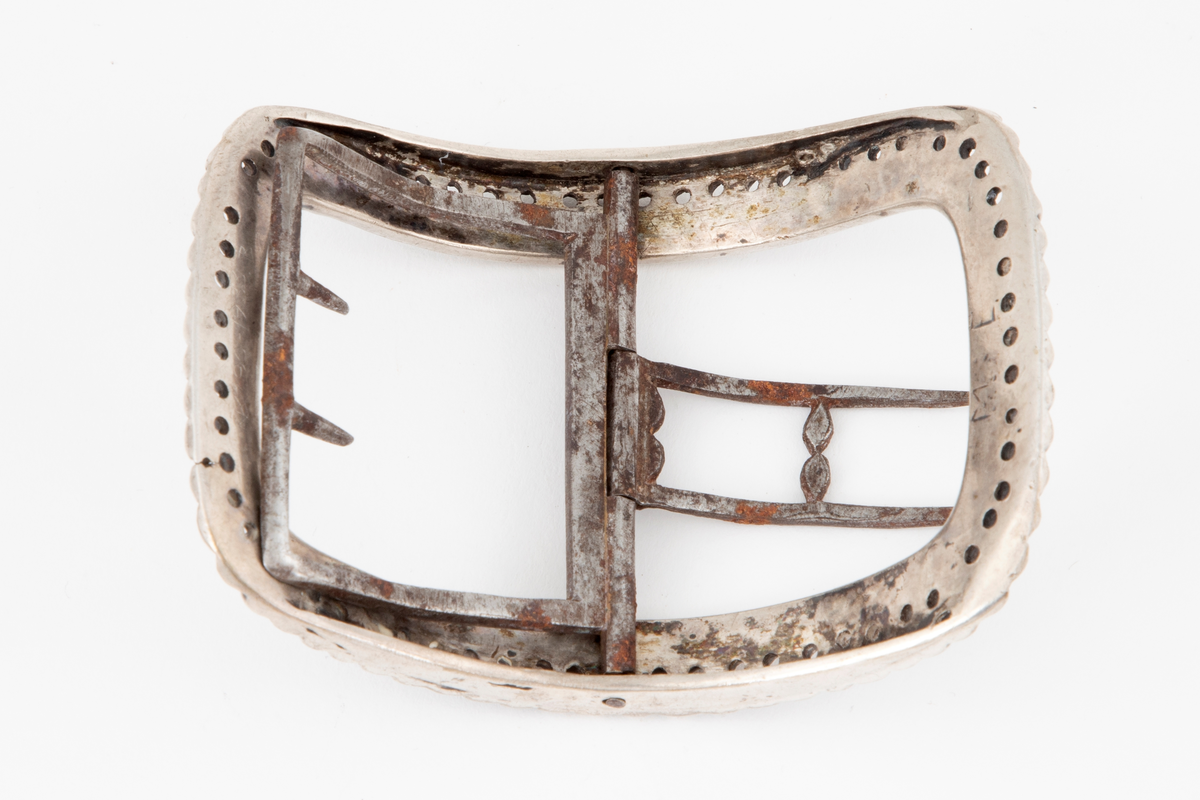 Skospenner støpt i sølv og siselert. Krummet, avlang rammeform med lett buede kortsider. Dekorert med to rader kantede "knopper", skilt med en rad små, gjennombrutte hull. Stolpe med dobbel torn i form av to parallelle tinner og feste med 2 små, faste torner, alt av jern.