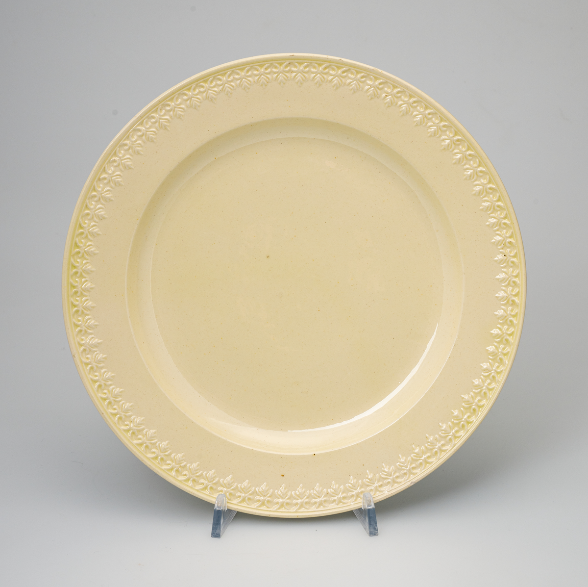 En flat tallerken av glasert steintøy (creamware). Den er hvitgul på farge (ensfarget). Flaten på midten er 16,5cm i diameter. Langs kanten er det tungekant. Innenfor tungekanten er det støpt dekor i form av en hjertebord med stiliserte blomster/bladverk. På undersiden er det dypstempel i form av fire små ruter/prikker i klase som er et pottemakermerke.