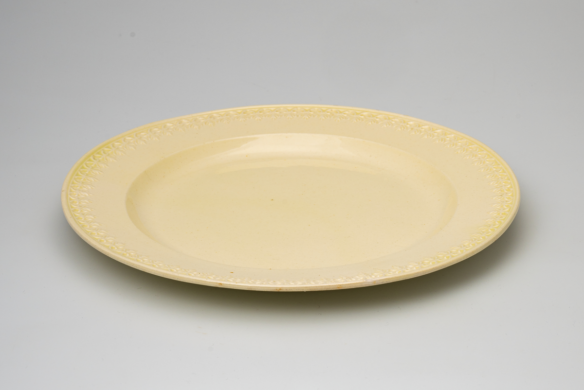 En flat tallerken av glasert steintøy (creamware). Den er hvitgul på farge (ensfarget). Flaten på midten er 16,5cm i diameter. Langs kanten er det tungekant. Innenfor tungekanten er det støpt dekor i form av en hjertebord med stiliserte blomster/bladverk. På undersiden er det dypstempel i form av fire små ruter/prikker i klase som er et pottemakermerke.