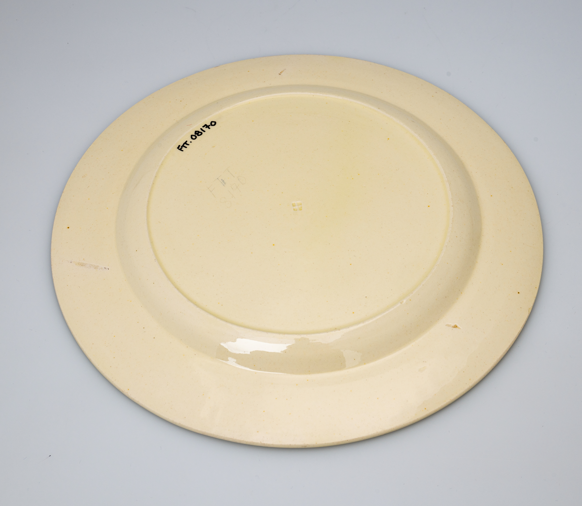 En flat tallerken av glasert steintøy (creamware). Den er hvitgul på farge (ensfarget). Flaten på midten er 16,5cm i diameter. Langs kanten er det tungekant. Innenfor tungekanten er det støpt dekor i form av en hjertebord med stiliserte blomster/bladverk. På undersiden er det dypstempel i form av fire små ruter/prikker i klase som er et pottemakermerke.