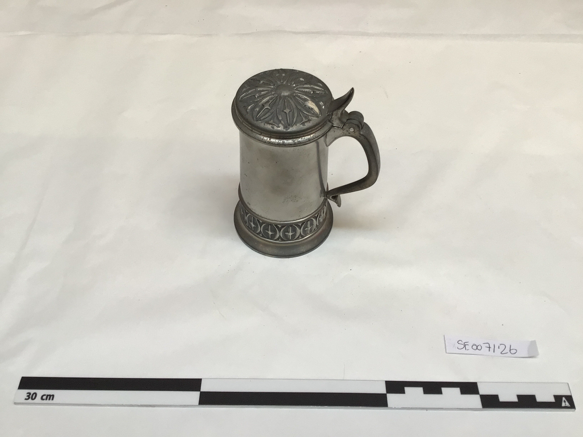 Drikkekar i tinn, rundt tverrsnitt, svakt kon. Hank i sida, hengsla lok og fotring. Loket er dekorert med ei åttebladrose, kvart roseblad har eit kors.  Det er eit band med korsmønster langsetter fotringen.