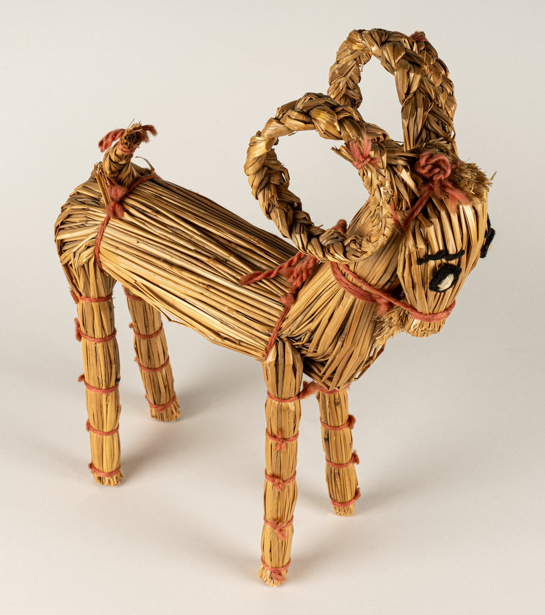 Julbock av halm, halmbock. Flätade horn och svans. Bocken är hopbunden med röda bomullsband. Ögon påsydda med svart och vitt garn.