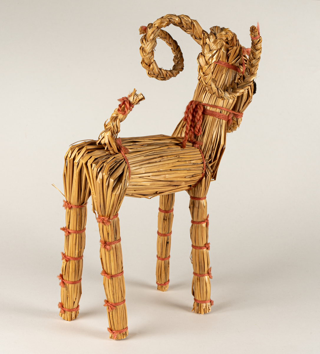 Julbock av halm, halmbock. Flätade horn och svans. Bocken är hopbunden med röda bomullsband. Ögon påsydda med svart och vitt garn.