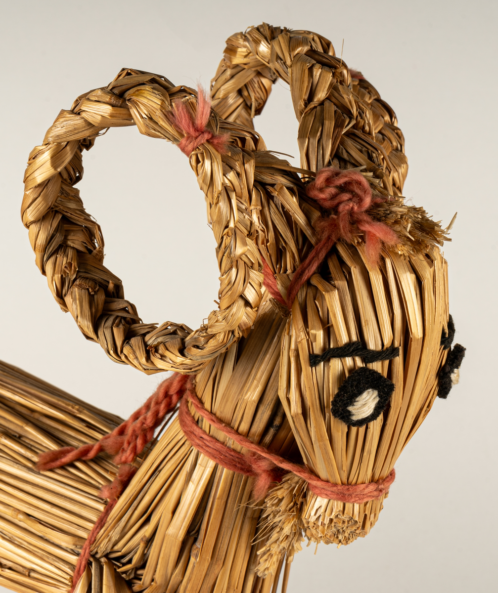 Julbock av halm, halmbock. Flätade horn och svans. Bocken är hopbunden med röda bomullsband. Ögon påsydda med svart och vitt garn.