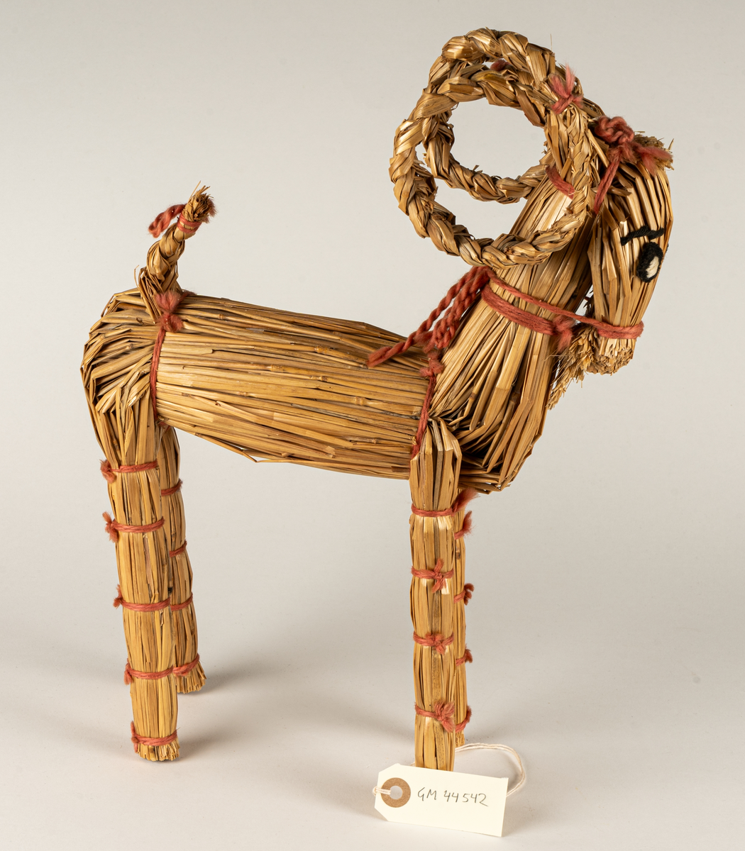 Julbock av halm, halmbock. Flätade horn och svans. Bocken är hopbunden med röda bomullsband. Ögon påsydda med svart och vitt garn.