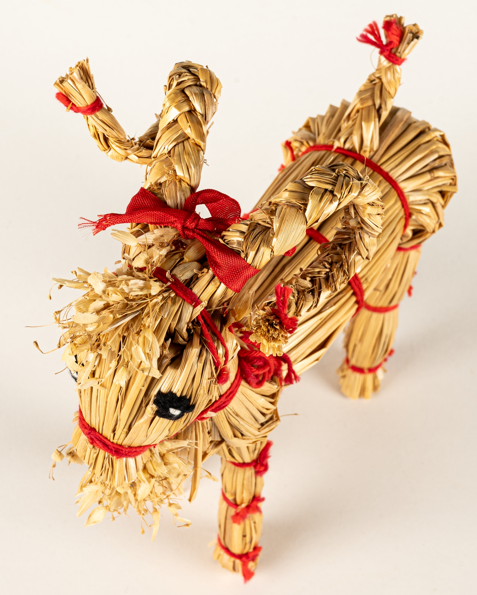 Julbock av halm, halmbock. Flätade horn och svans. Bocken är hopbunden med röda bomullsband. Ögon påsydda med svart och vitt garn.