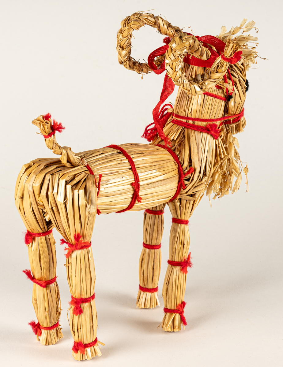 Julbock av halm, halmbock. Flätade horn och svans. Bocken är hopbunden med röda bomullsband. Ögon påsydda med svart och vitt garn.