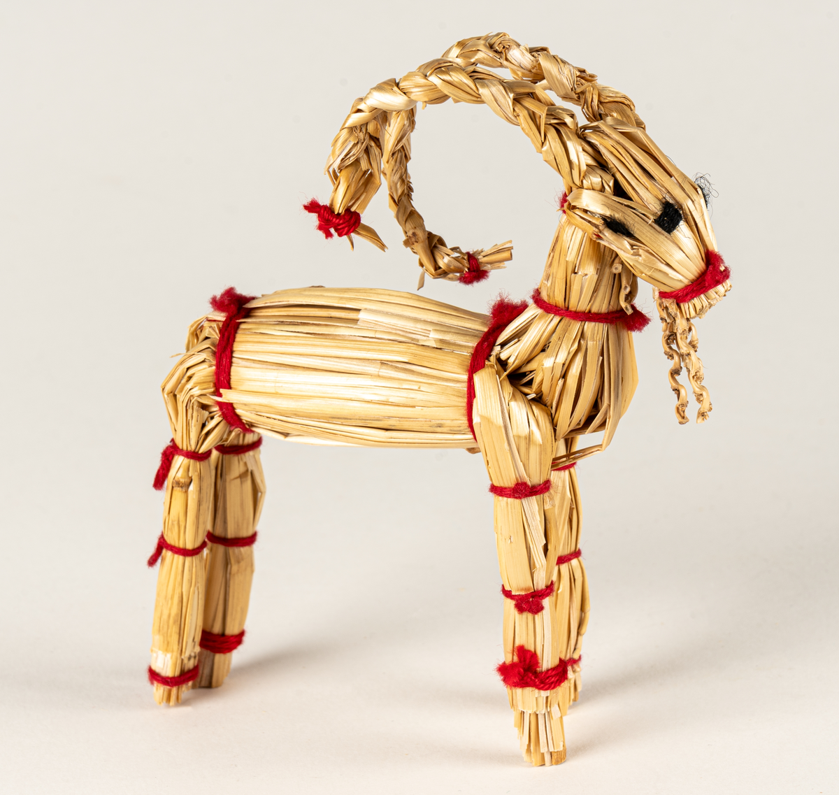 Julbock av halm, halmbock. Flätade horn och svans. Bocken är hopbunden med röda bomullsband. Ögon påsydda med svart och vitt garn.