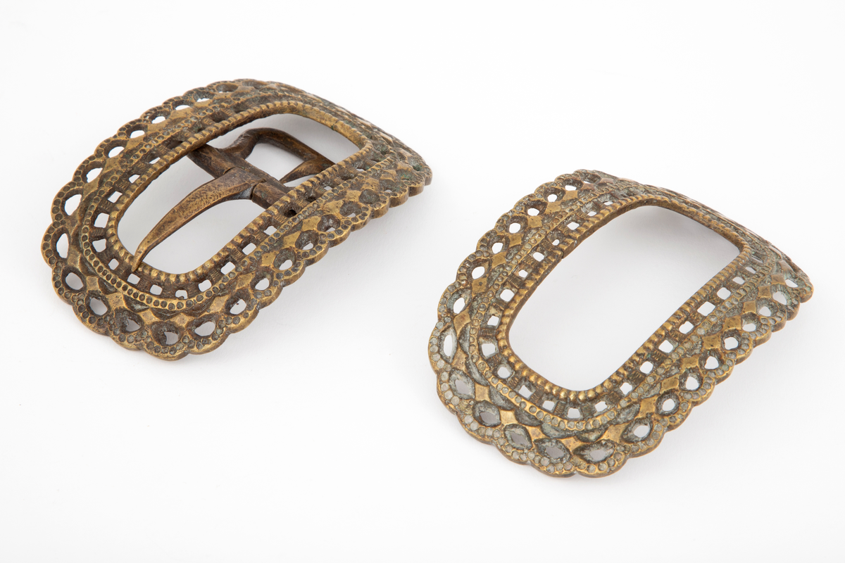 Skospenner støpt i krummet, oval form med tilsvarende innvendig åpning. Gjennombrutt med perlerader, groper, hull og ytterst tunget, gjennombrutt rand. Kun den ene spennen har stolpe med torn og feste med liten fast torn.