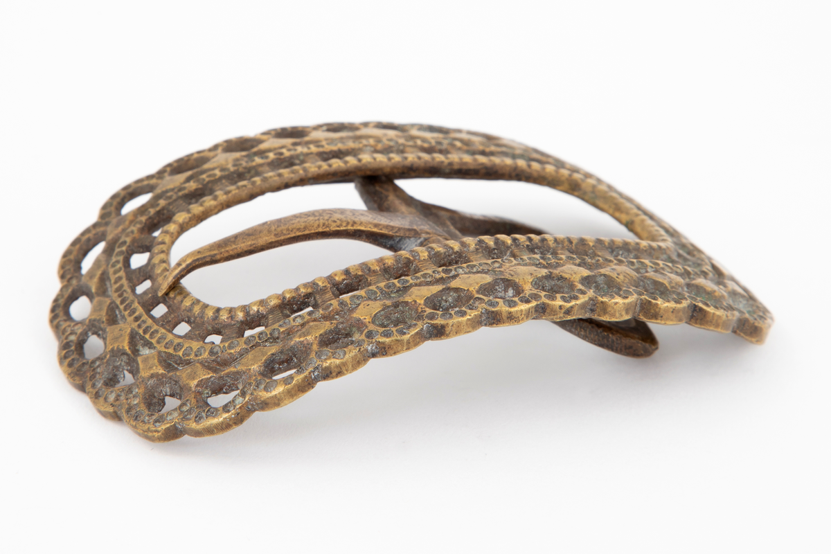 Skospenner støpt i krummet, oval form med tilsvarende innvendig åpning. Gjennombrutt med perlerader, groper, hull og ytterst tunget, gjennombrutt rand. Kun den ene spennen har stolpe med torn og feste med liten fast torn.