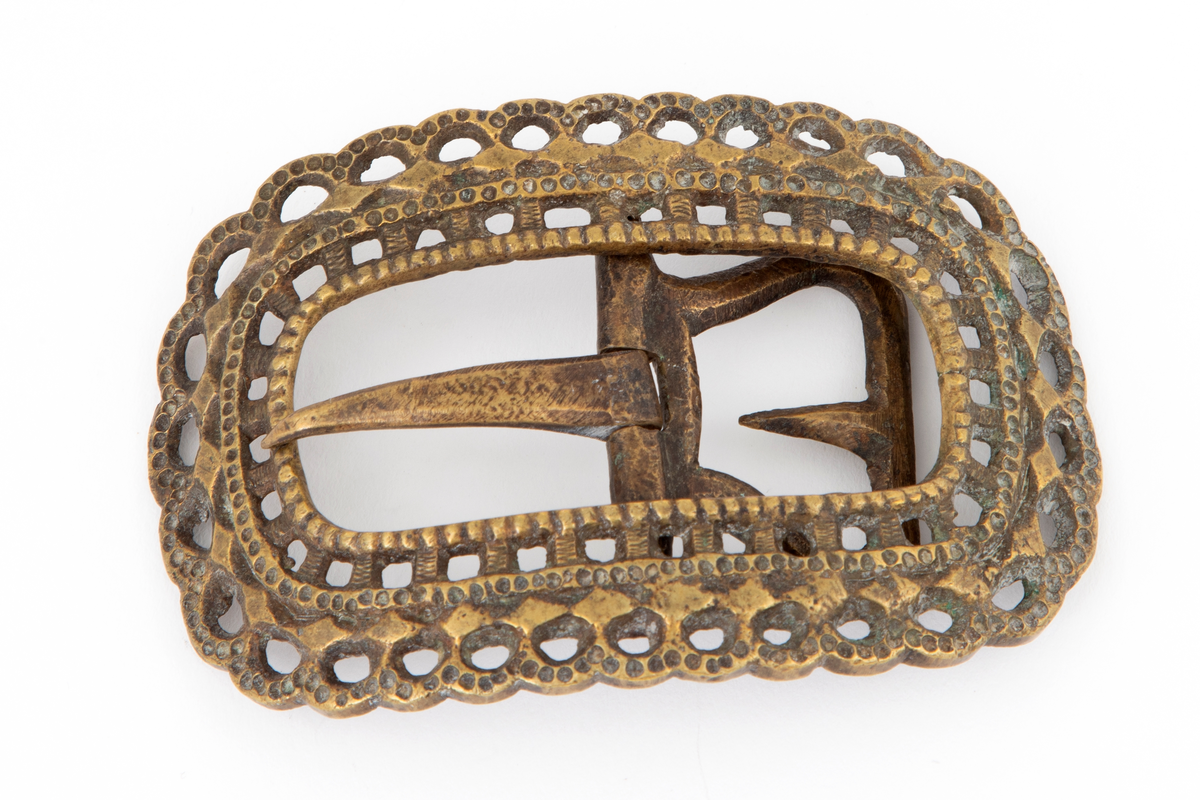 Skospenner støpt i krummet, oval form med tilsvarende innvendig åpning. Gjennombrutt med perlerader, groper, hull og ytterst tunget, gjennombrutt rand. Kun den ene spennen har stolpe med torn og feste med liten fast torn.