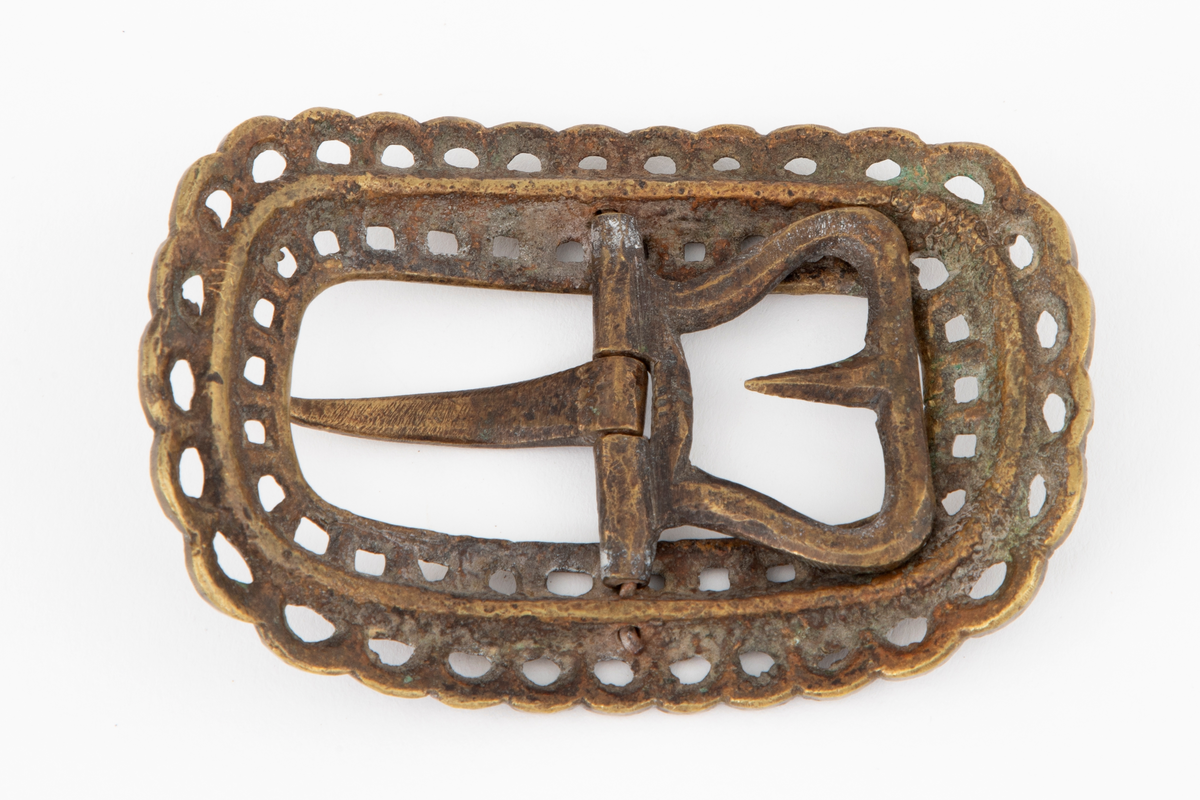 Skospenner støpt i krummet, oval form med tilsvarende innvendig åpning. Gjennombrutt med perlerader, groper, hull og ytterst tunget, gjennombrutt rand. Kun den ene spennen har stolpe med torn og feste med liten fast torn.