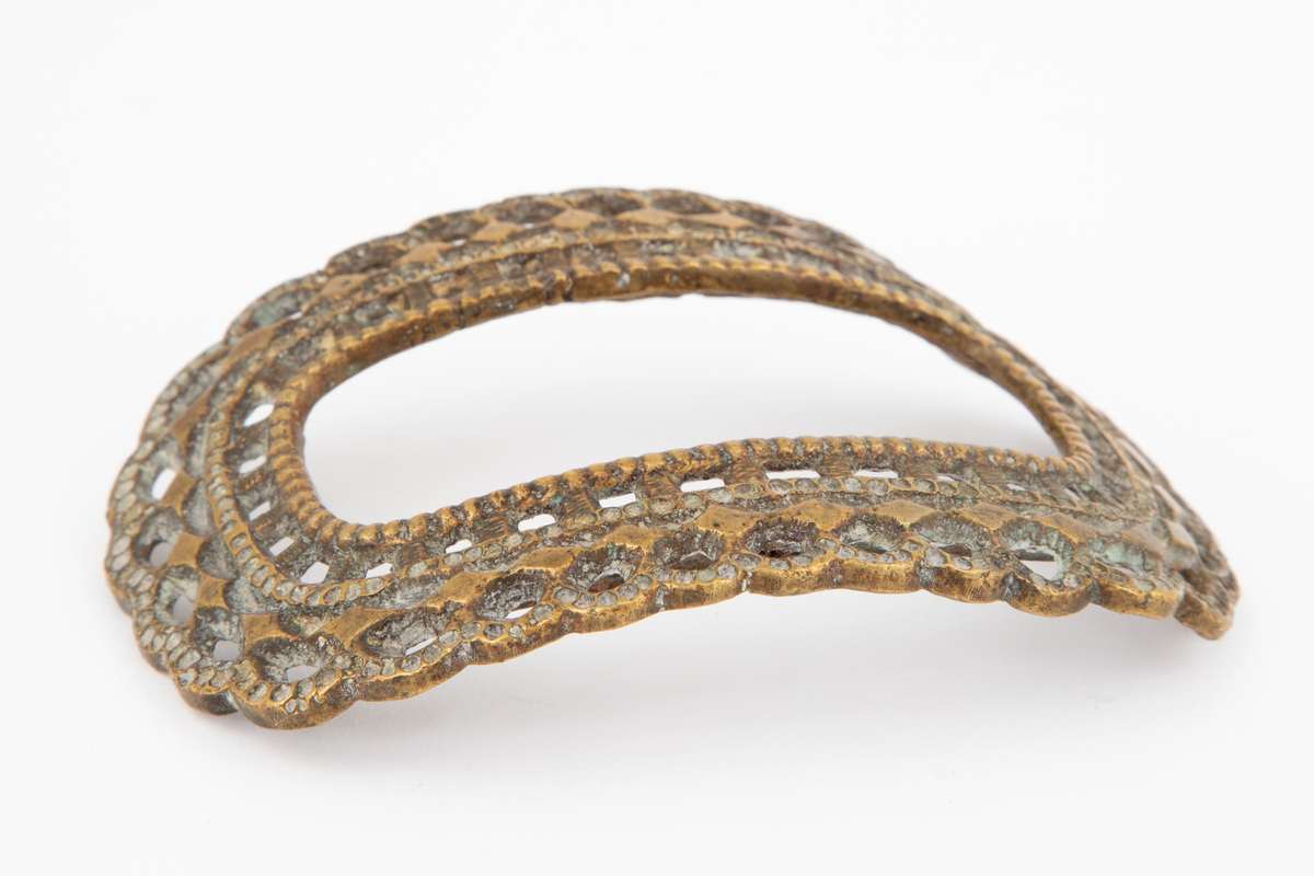 Skospenner støpt i krummet, oval form med tilsvarende innvendig åpning. Gjennombrutt med perlerader, groper, hull og ytterst tunget, gjennombrutt rand. Kun den ene spennen har stolpe med torn og feste med liten fast torn.