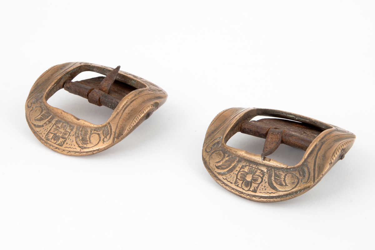 Skospenner av bronse støpt i krummet, nesten rund form med indre firkantet åpning. På to av rammesidene siselert rosett og volutter. De to andre sidene har båndlignende ornament. Stolpe med torn og feste i form av tunge med knapp i jern.