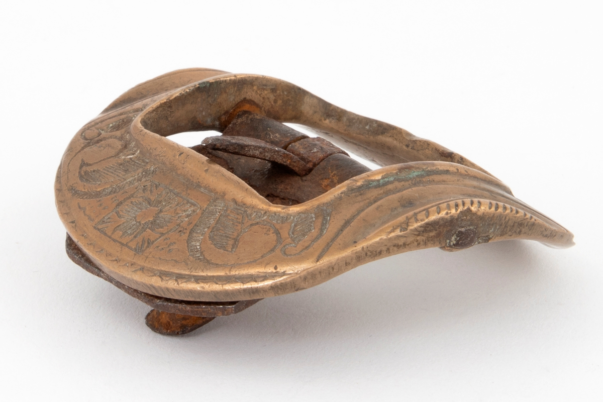 Skospenner av bronse støpt i krummet, nesten rund form med indre firkantet åpning. På to av rammesidene siselert rosett og volutter. De to andre sidene har båndlignende ornament. Stolpe med torn og feste i form av tunge med knapp i jern.