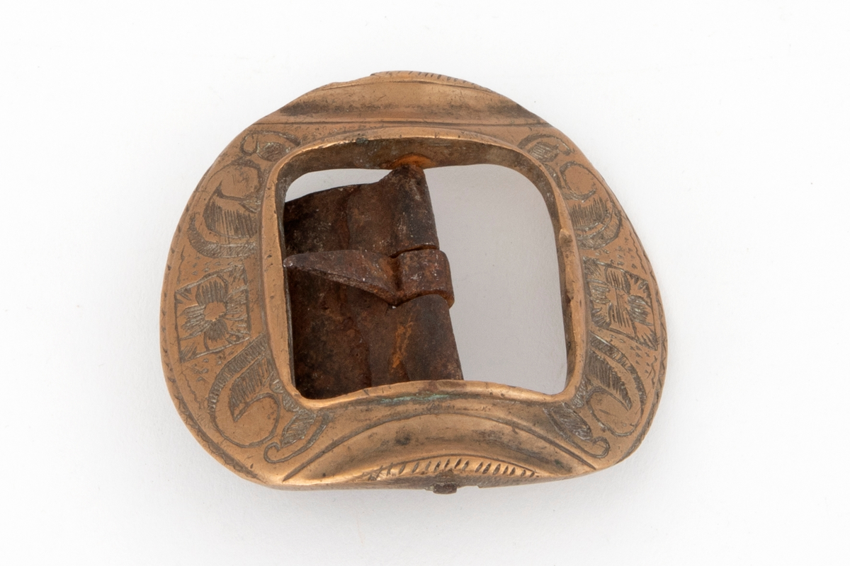 Skospenner av bronse støpt i krummet, nesten rund form med indre firkantet åpning. På to av rammesidene siselert rosett og volutter. De to andre sidene har båndlignende ornament. Stolpe med torn og feste i form av tunge med knapp i jern.