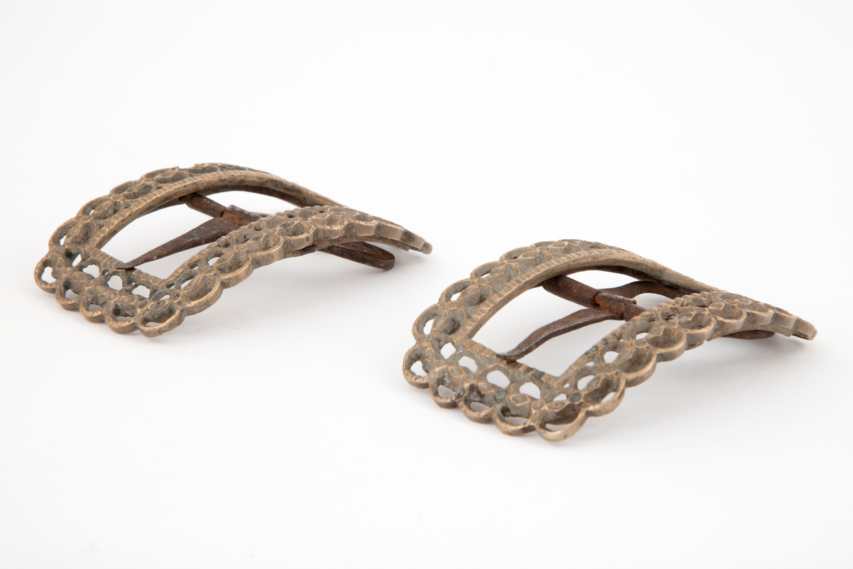 Skospenner av bronse støpt i krummet, rektangulær form med avrundede hjørner. Indre rektangulær åpning med skarpe hjørner. Rammen er gjennombrutt med dobbelt rad motstilte buer. Mellom buene bord med rombiske og runde groper. Stolpe med torn og feste med liten fast torn i jern.